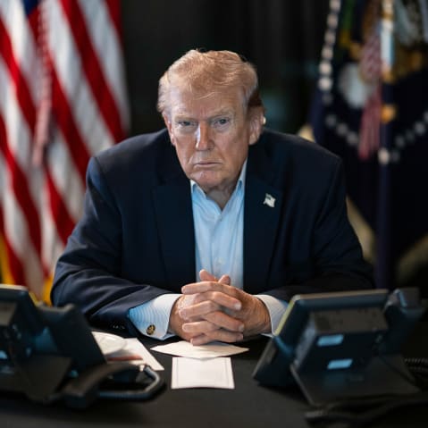 Donald Trump sits at a table with two phones in front of him and U.S. flags behind him.