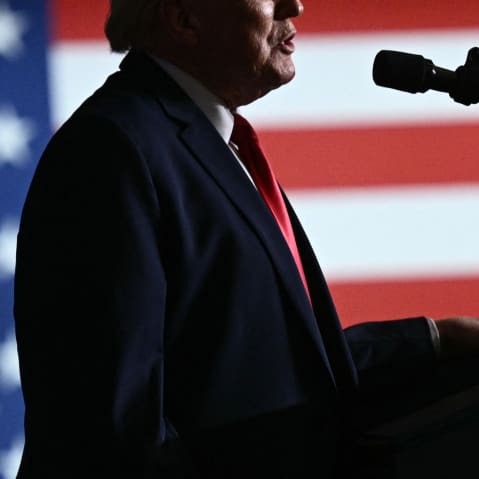 Donald Trump's mouth and torso in profile speaking into a microphone. Behind him, the American flag.