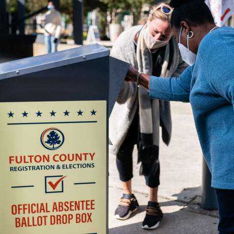 A woman helps another woman to put her absentee ballot into a box outside.