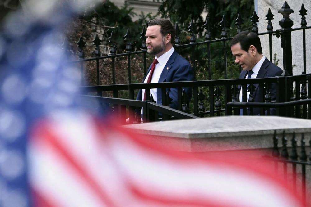 Out of focus in the foreground is the American flag, occupying the left and bottom of the frame. JD Vance, left, and Marco Rubio are in focus in the background, seen walking out of a building.