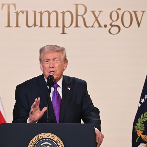 Donald Trump speaks into a microphone at a podium. The website "TrumpRx.gov" is on the wall behind him.
