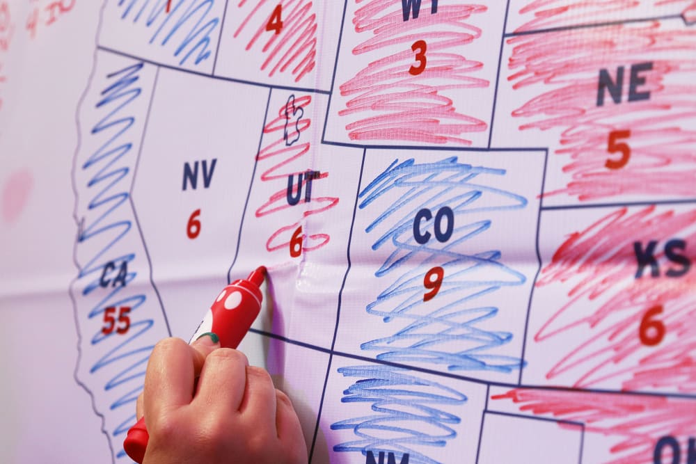 A hand holding a red marker colors Utah red on a map. Some of the other states have already been colored in either red or blue; others are still blank.