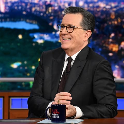 Stephen Colbert laughs while he sits at his desk.
