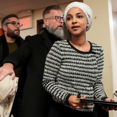 Ilhan Omar walks into a room for a town hall meeting, followed by two men who seem to be security personnel.