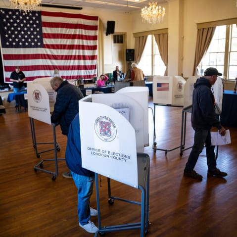 People vote in a polling location with a large American flag in the background.