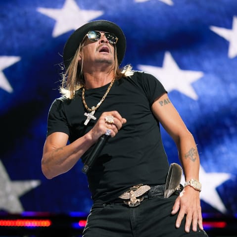 Kid Rock holds a microphone on stage. The background is an American flag.
