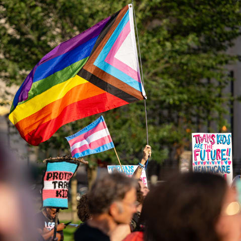 People hold signs in support of trans kids and LGBTQ+ flags.