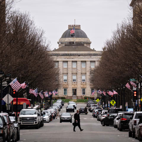 A street lined with cars leads up to the North Carolina State Capitol. A person crosses in the middle of the street, and American flags are on each of the lampposts.