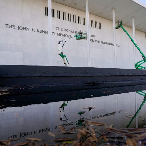 Workers in green construction lifts work on the facade of the Kennedy Center, which is reflected in a puddle.