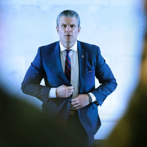 Pete Hegseth walks toward the camera behind two people who arew out of focus in the foreground.