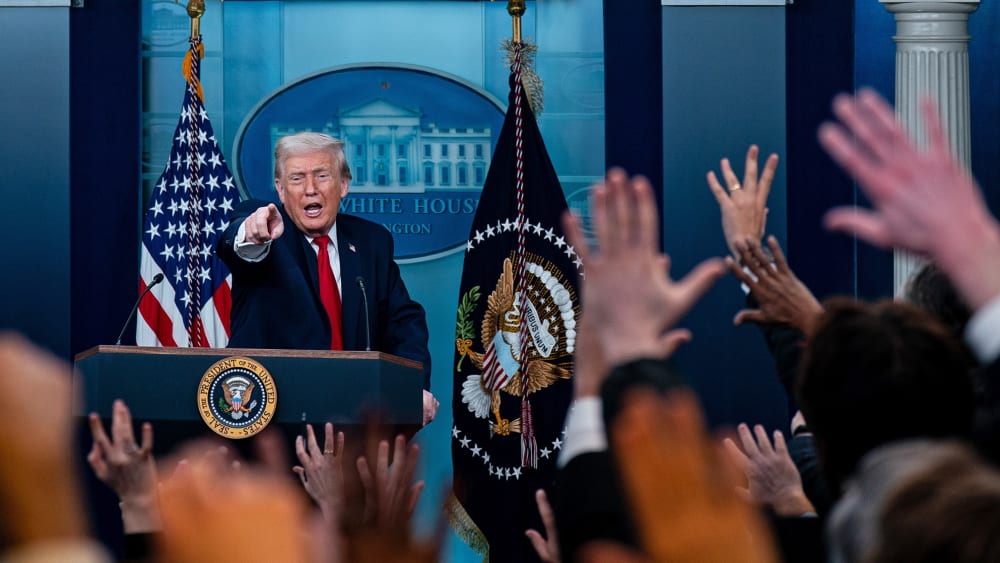 Donald Trump, in focus, stands a podium and points in front of him. Out of focus in the foreground, many people raise their hand.