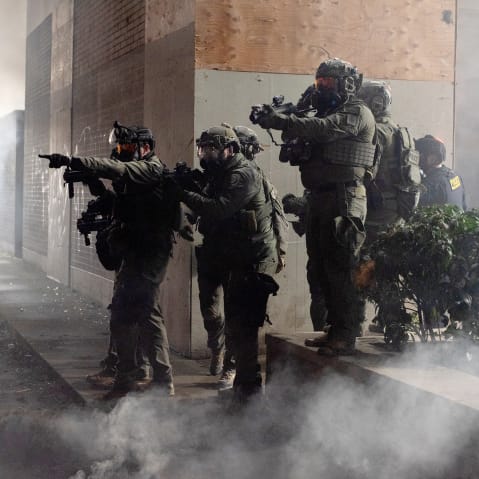 Federal agents are seen in a cloud of smoke in tactical gear, targeting protestors with their weapons.