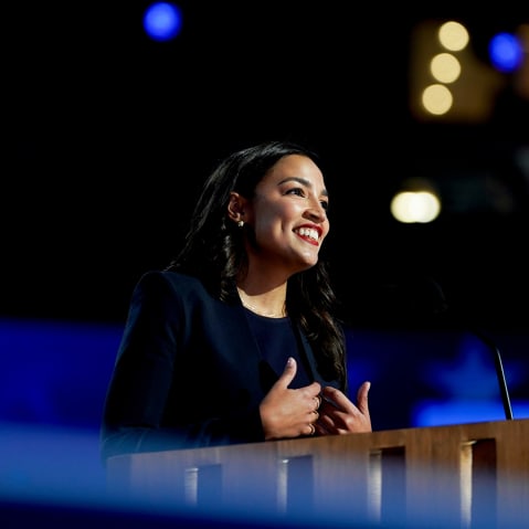 Alexandria Ocasio-Cortez smiles while standing behind a podium.