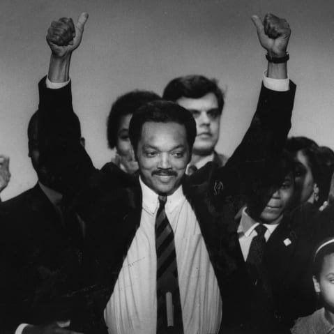 Jesse Jackson Sr. lifts his arms surrounded by a group of people mixed-aged people.