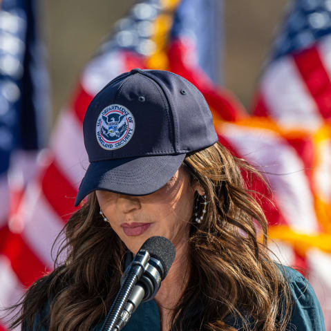 Kristi Noem with her head tilted down, her "U.S. Department of Homeland Security" baseball cap obscuring her eyes.