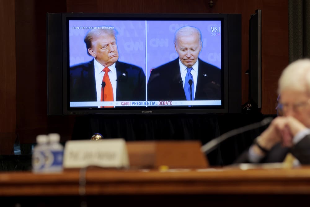 Donald Trump and Joe Biden are seen on a split screen while there is a man sitting in front of the screen to the right of the frame, out of focus.