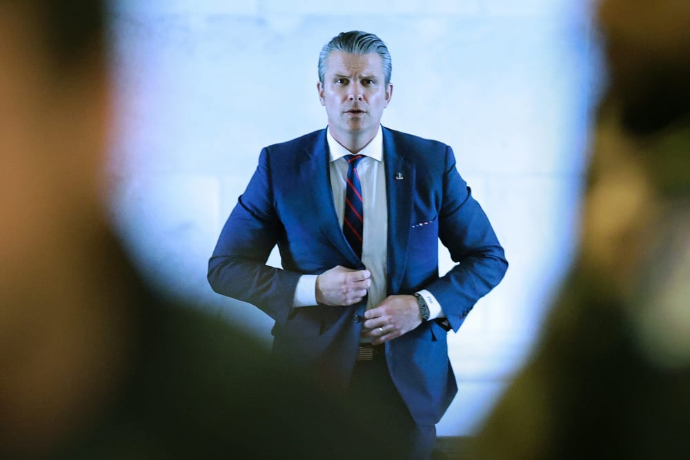 Pete Hegseth walks toward the camera behind two people who arew out of focus in the foreground.