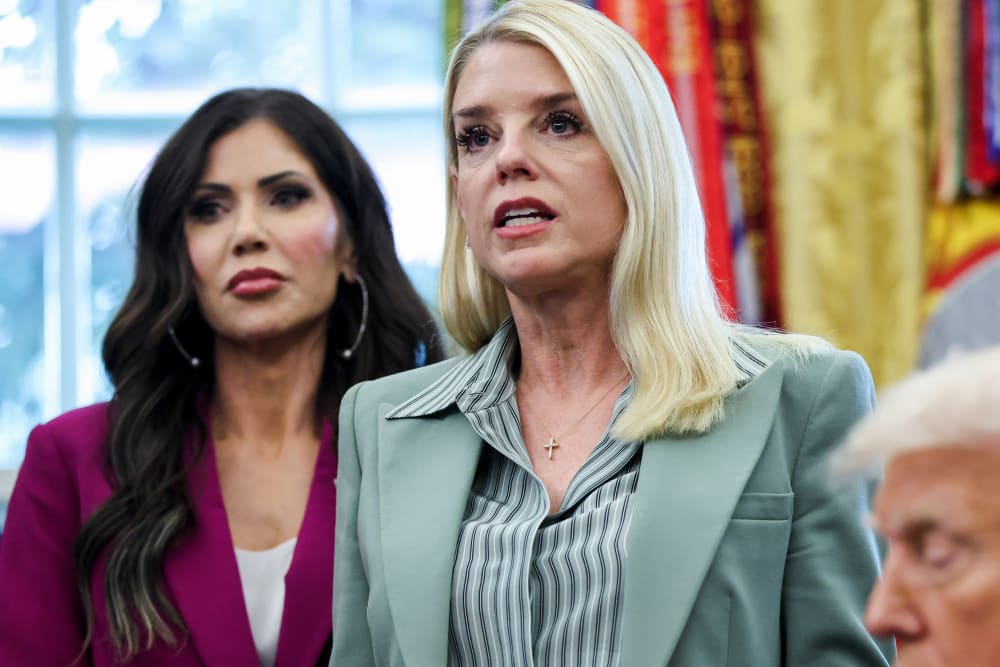 Kristi Noem is seen on the left side of the frame as she stands behind Pam Bondi. A small part of Donald Trump's face is visible on the bottom right.
