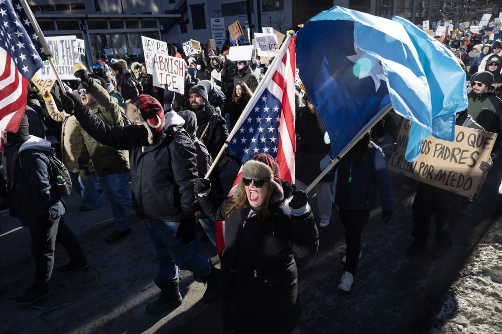 A female protestor shouts as she carries a flag in the foreground where others follow her during a daytime demonstration.