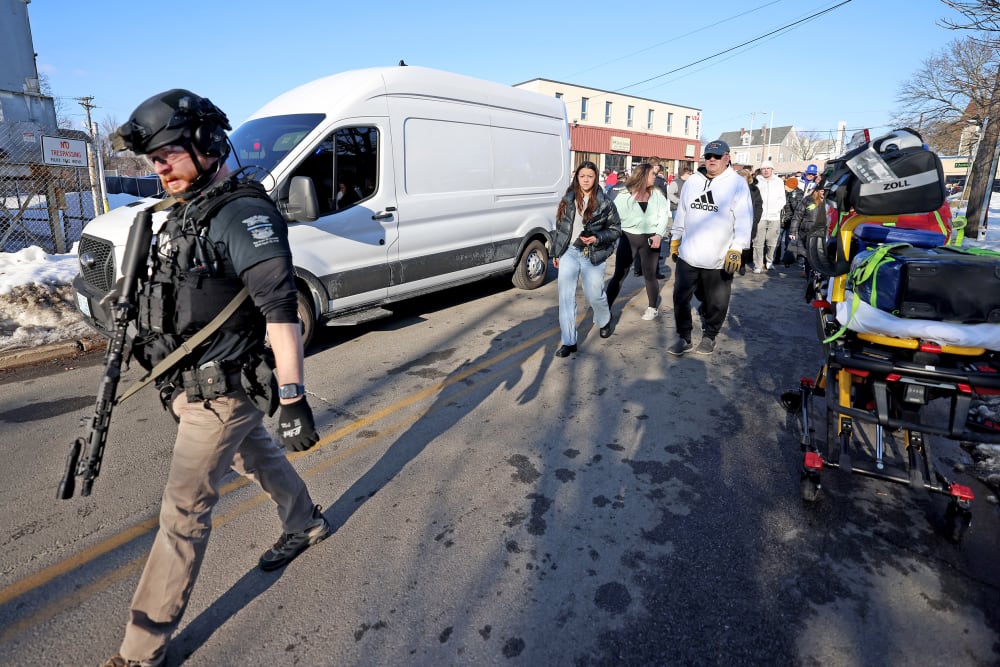 An armed police officer walks as other civilian people follow him.