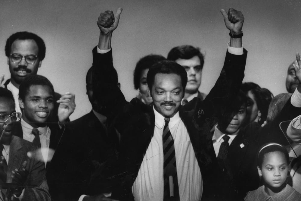 Jesse Jackson Sr. lifts his arms surrounded by a group of people mixed-aged people.
