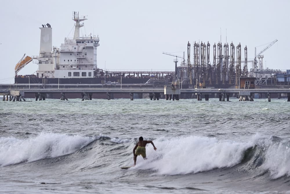 A man is seen from behind as he surfs in front of an oil tanker in the distance.