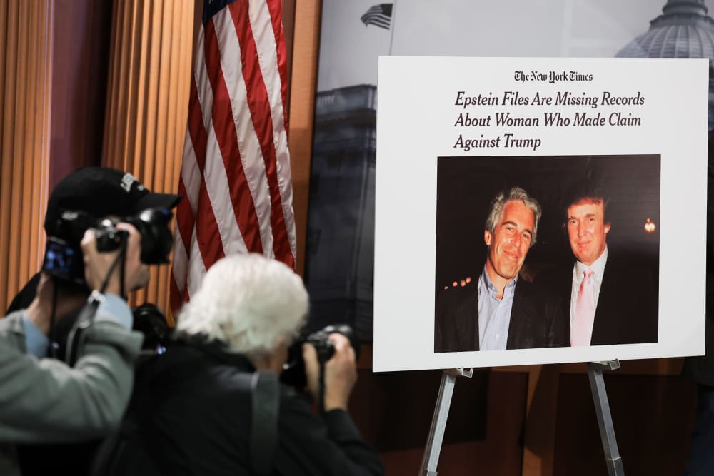 There are two photographers on the left bottom of the frame taking pictures of a poster that shows a photo of Donlad Trump and Jeffrey Epstein together.