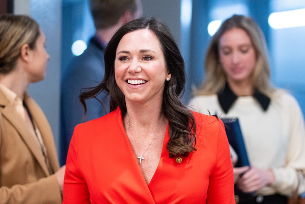 Katie Britt walks toward the camera with a smile on her face, wearing a red jacket.