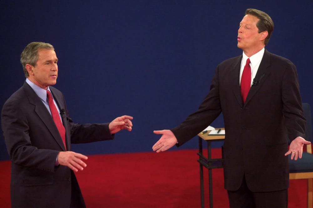 George Bush speaks to Al Gore while Gore reacts by opening both arms.
