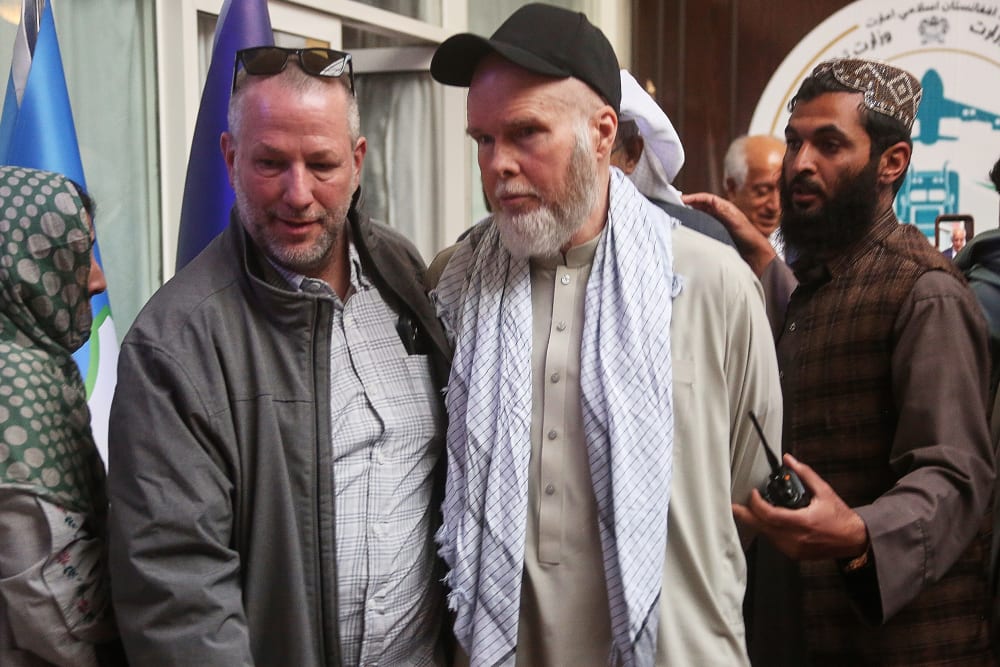 Dennis Coyle looks away, wearing a traditional Afghani clothing with a baseball cap as her is escorted by two men.
