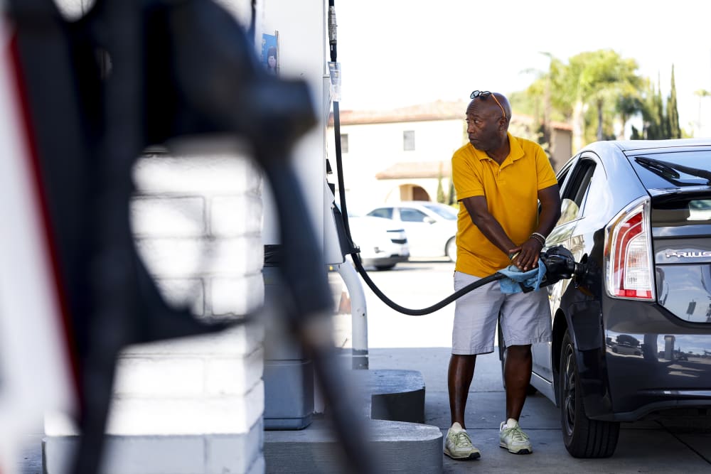 A man in a yellow T-shirt pumps gas on the right side of the frame. We can see another gas pump in the foreground.