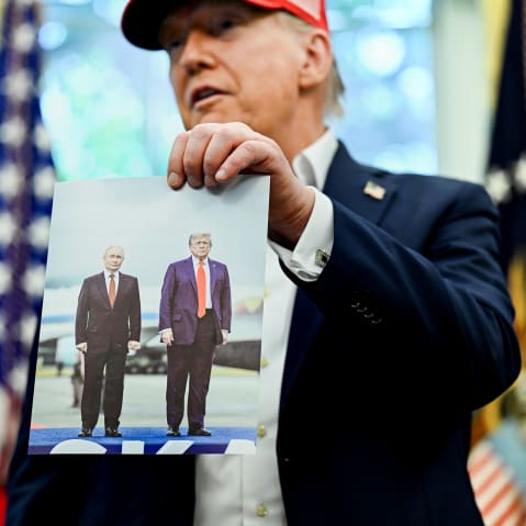 President Trump holds photo of him and Putin in Alaska while he is wearing a red cap in the oval office.