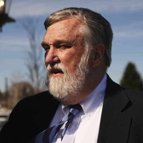 Doug Wilson stands next to a wall facing the sun, his eyes are squinted.