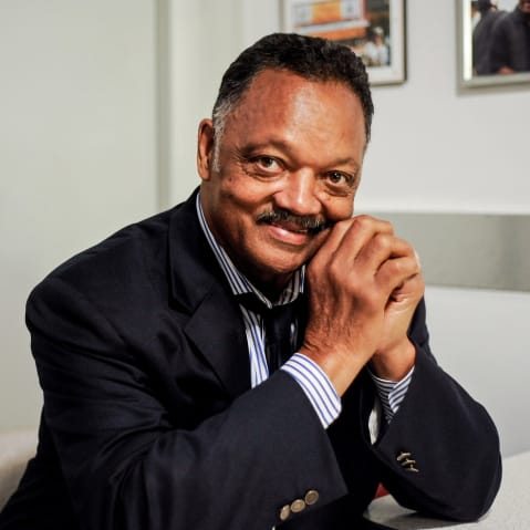 Rev. Jesse Jackson poses for a portrait, sitting down with his hands clasped up by his face.