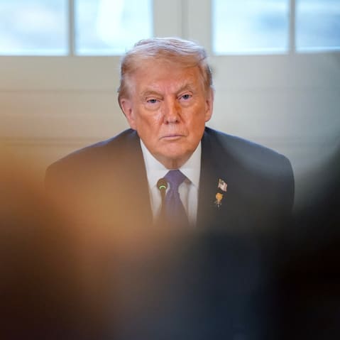 Donald Trump looks on, partially obscured by people in the foreground.