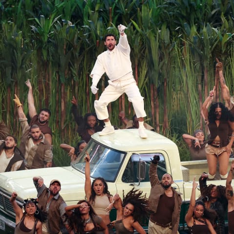 Bad Bunny stands on the roof of a light yellow truck. People dance in the truck bed and on the football field below.