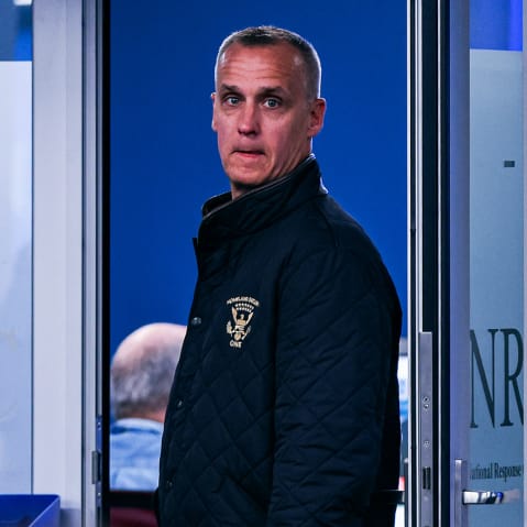 Corey Lewandowski stands in a doorway. Blurred in the foreground, people work at their desks.