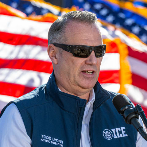 Todd Lyons speaks at a podium outside during daytime.