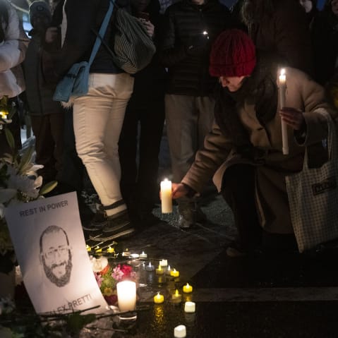 A person kneels down to place a lit candle by a memorial featuring an illustration of Alex Pretti.
