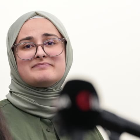 Rumeysa Ozturk looks at the woman speaking at the podium.