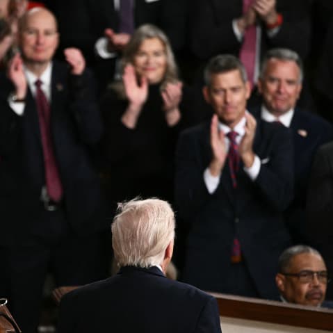 People clap, out of focus, as the back of Donald Trump's head is seen in focus in the foreground.