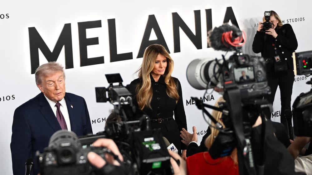 Donald and Melania Trump stand in front of a step-and-repeat banner advertising the movie "Melania." In front of them are journalists with cameras.