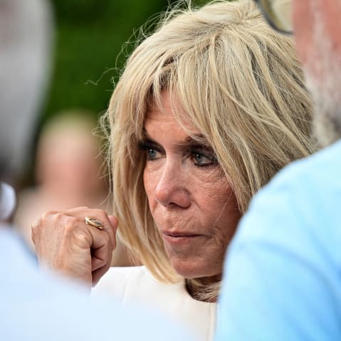 Brigitte Macron, right, looks on while standing with two males. This is a closeup frame.