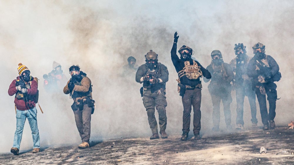 One federal agent, walking through a cloud of tear gas, throws a teargas canister which is now suspended in midair. Other federal agents walk alongside.