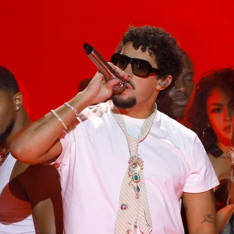 Bad Bunny, surrounded by performers, holds a microphone against a red background.