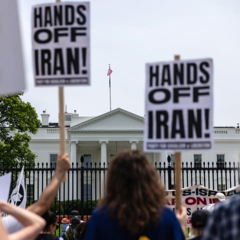 Out of focus in the foreground, people hold picket signs that read "Hands Off Iran!" In focus in the background is the White House.