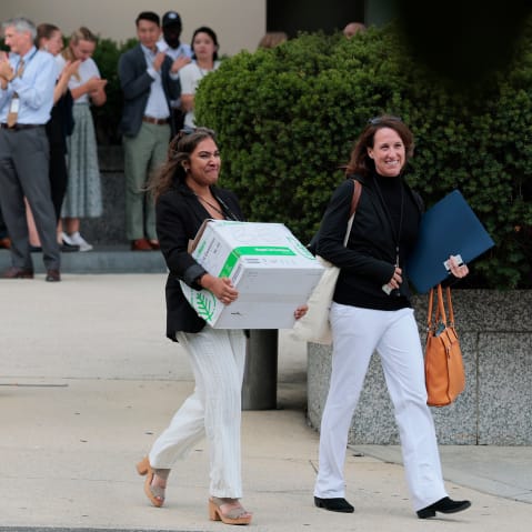 People carrying boxes of stuff walk into a parking lot; people behind them clap.
