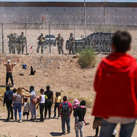 Adults and children with their backs to the camera look up at people in uniform who are behind a barbed wire fence.