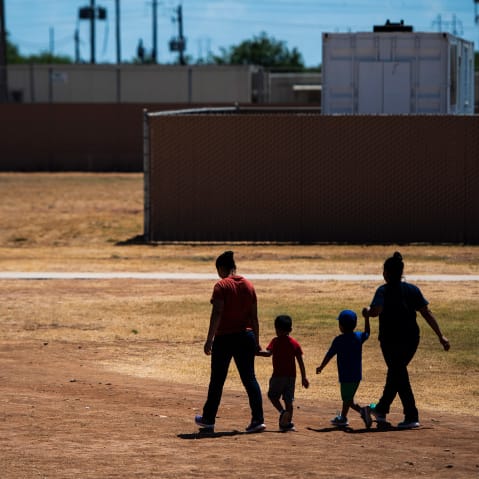 Two women, each holding the hand of a child, walk across a field with their backs to the camera.