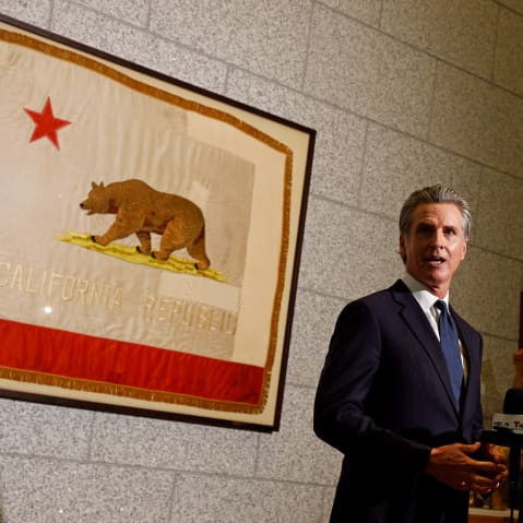 Gavin Newsom stands behind news microphones and in front of a framed California state flag.